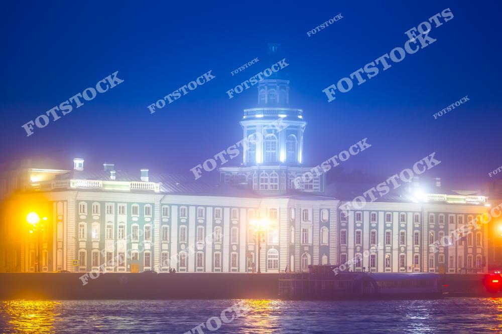 На фотографии изображена Кунсткамера, известный музей антропологии и этнографии, расположенный на Васильевском острове в Санкт-Петербурге. Ее характерная архитектура и яркое освещение делают здание особенно привлекательным в условиях тумана. Уличные фонари освещают набережную и придают особый шарм этому историческому месту. Этот вид символизирует культурное наследие Петербурга и его уникальную атмосферу.